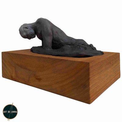 A photo of a sculpture titled "Grief."It is a clay sculpture portraying a man's battle against the relentless tide of sorrow. The weight of his exhaustion as he emerges from it's depth, his form painted in a subtle ombre from dark charcoal to light grey. Mounted on a solid 50mm Kiaat timber block, this 20x15x13cm sculpture invites contemplation of the universal human experience of loss and resilience.